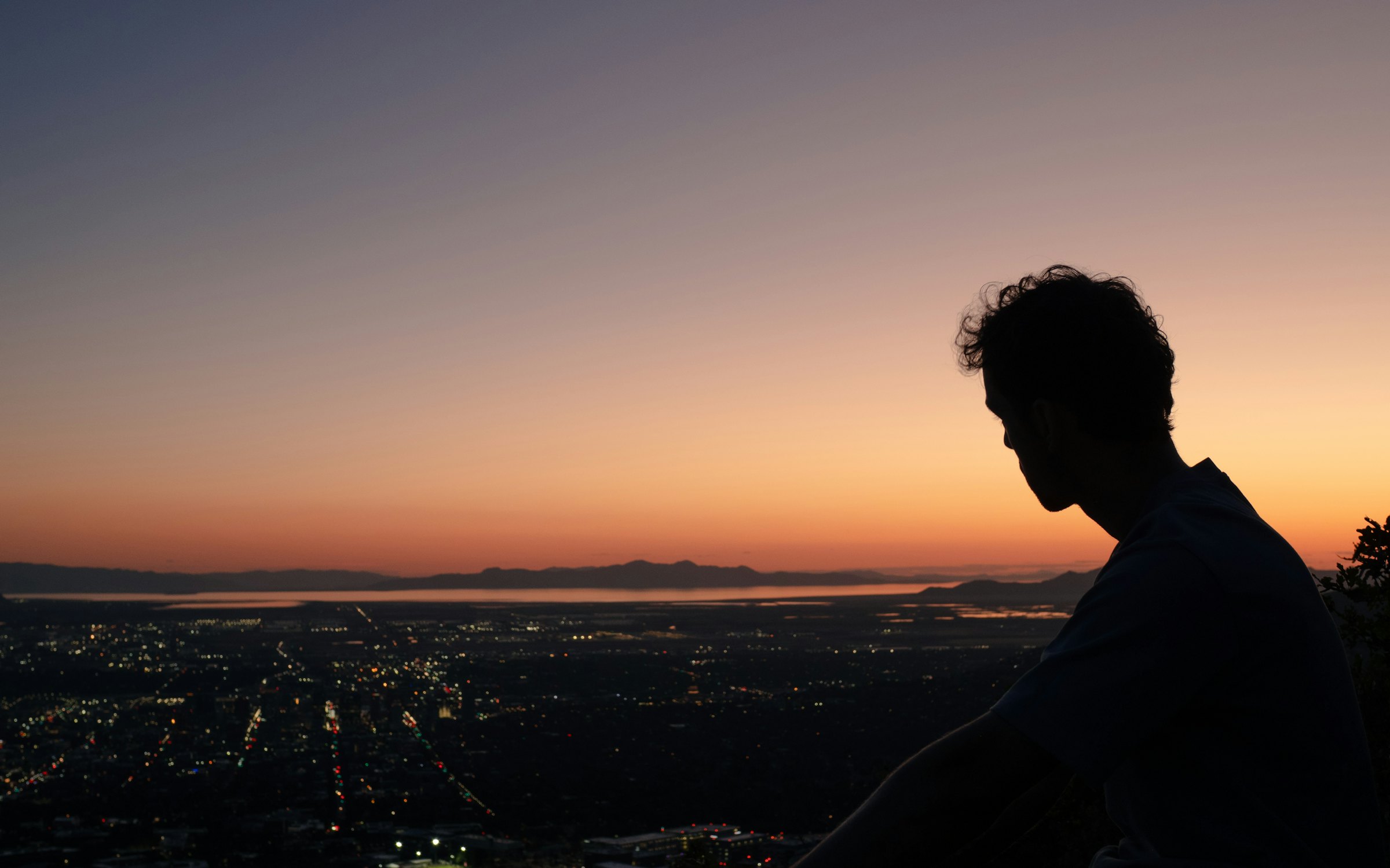 Um jovem, em silhueta, olha do alto de um morro as luzes de uma cidade no anoitecer.